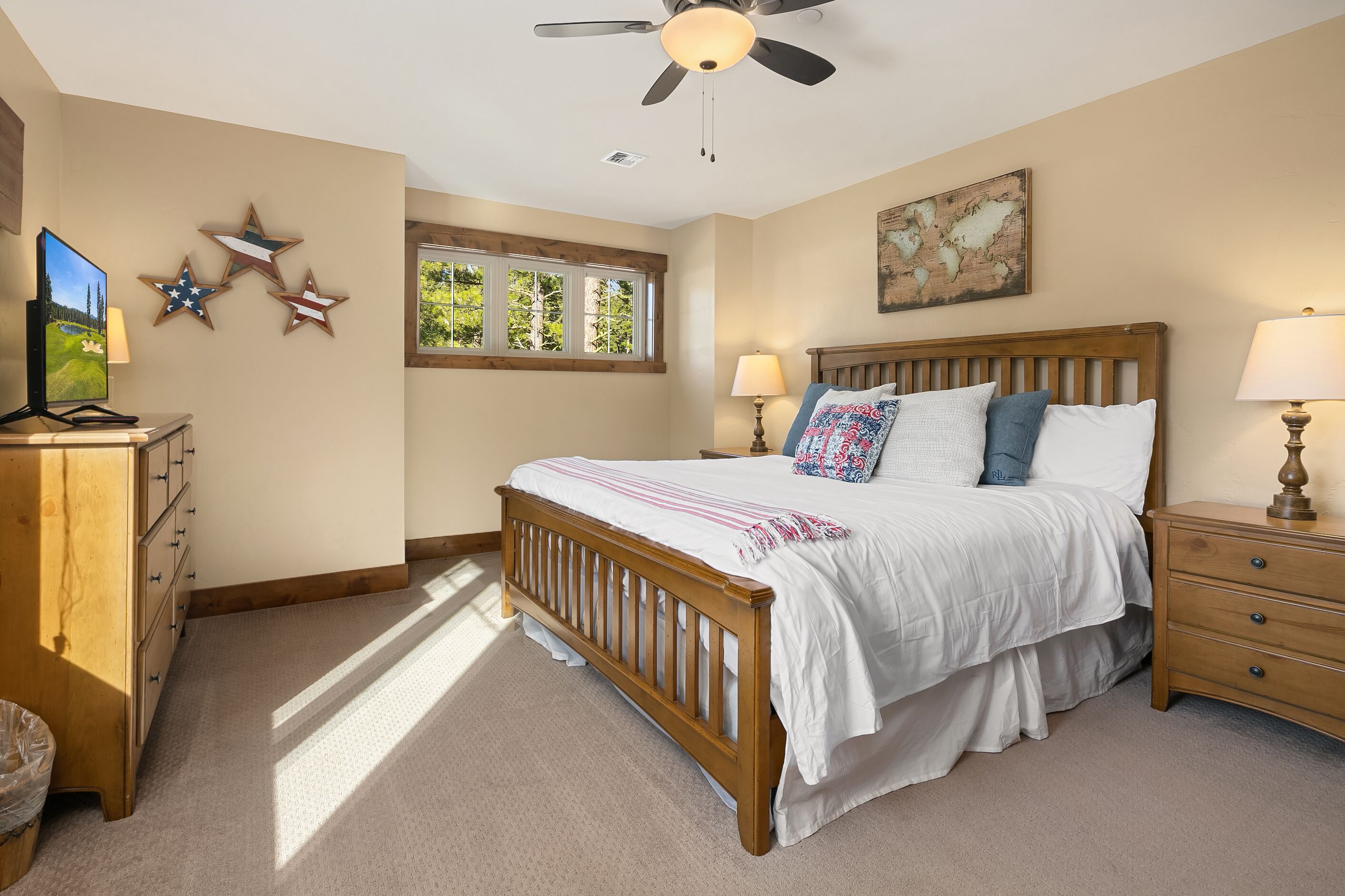 Un dormitorio con una cama de madera, dos mesitas de noche, un ventilador de techo, un ventanal con vistas a árboles y decoraciones en la pared que incluyen estrellas y un mapa.