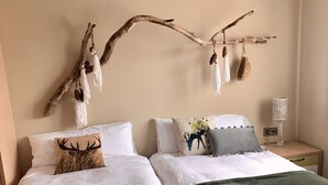 Un dormitorio con dos camas, ropa de cama blanca, una decoración de ramas con objetos colgantes y una mesita de noche con una lámpara.