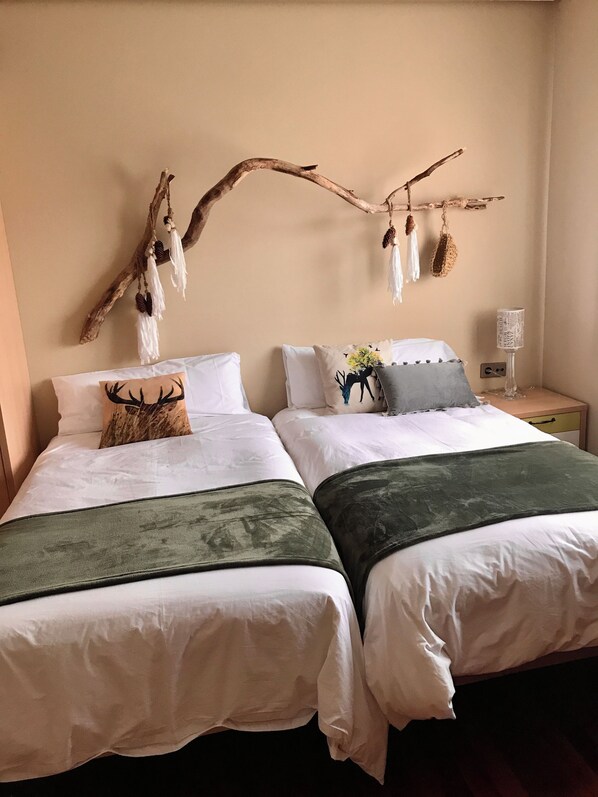 Quarto com duas camas, roupa de cama branca, um adorno de galho com objetos pendurados e uma mesa de cabeceira com abajur.