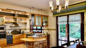 Una cocina con armarios de madera, una isla central con taburetes, una mesa de comedor con sillas y vistas a la vegetación a través de amplios ventanales.