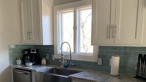 Una cocina con armarios blancos, encimera de granito, pileta de acero inoxidable y ventana con vista a árboles.