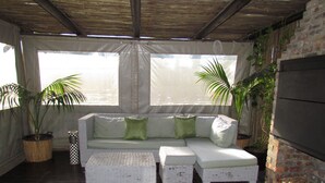 A covered outdoor seating area with a wicker sofa and cushions, surrounded by potted plants and large windows. - Princess Vacation Home (Richwood)