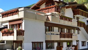 Un immeuble à plusieurs étages doté de balcons en bois, d’une façade en pierre et d’un espace extérieur aménagé pour s’asseoir.