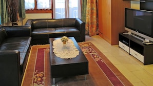 A living room with a black leather sofa, a coffee table with a lace doily, a flat-screen TV on a stand, and a wooden cabinet.
