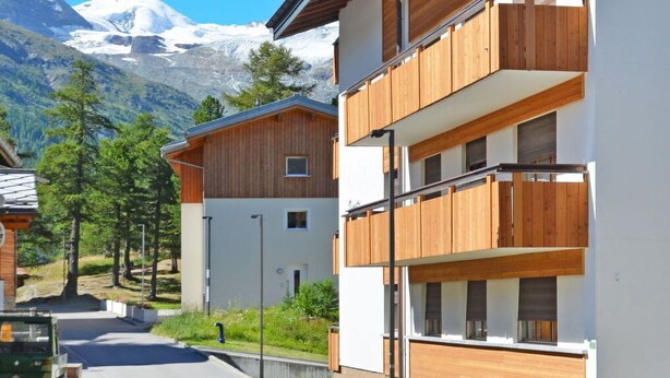 A modern multi-story building with wooden balconies, set against a backdrop of a snow-capped mountain and clear blue sky.