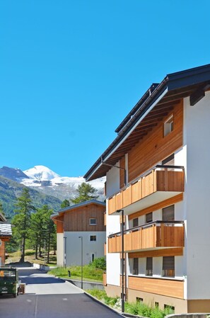 A modern multi-story building with wooden balconies, set against a backdrop of a snow-capped mountain and clear blue sky. - Lovely apartment for 5 people with WIFI, TV and terrace (Saas-fee)