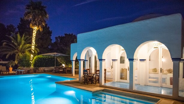 A swimming pool with a clear blue water surface, surrounded by a building with arched doorways and a patio area with chairs and umbrellas. - Villa, 8BR, Sea views, walk to the beach, pool house (Ibiza)