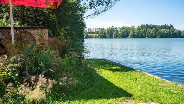 A lakeside view with a grassy area, a stone structure, and a red banner. - Beautiful private villa for 6 guests with WIFI, A/C, TV, patio and pets allowed (Malý Ratmírov)