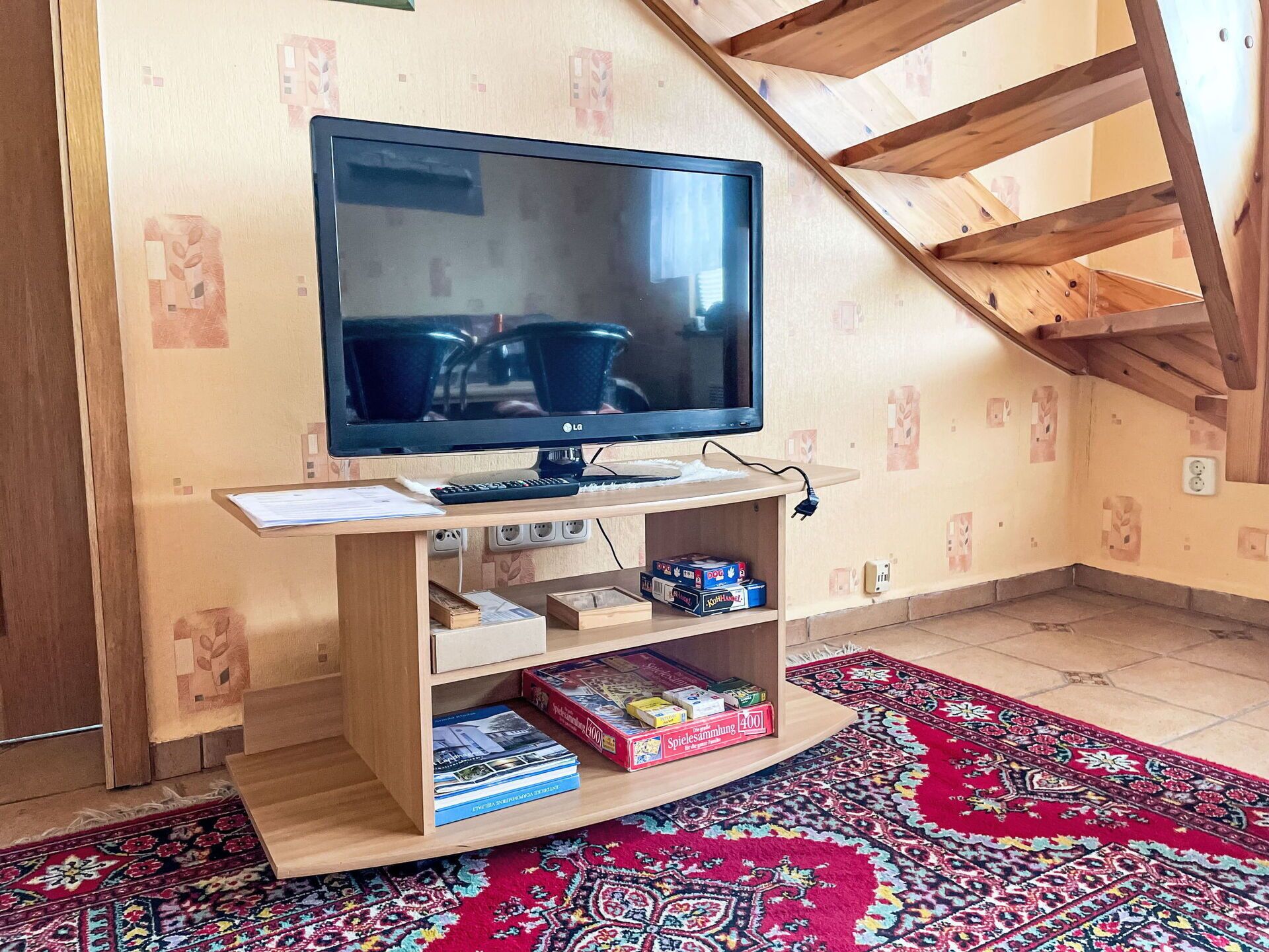 A television on a wooden stand with books and a remote control, a patterned rug, and a wooden ceiling structure.