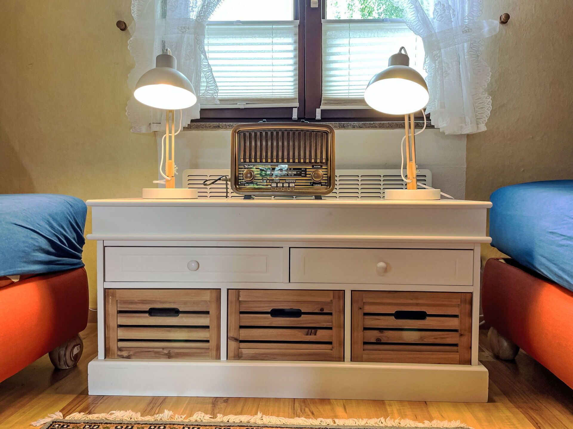 A room with a white bedside table, two lamps, a vintage radio, and two orange beds.