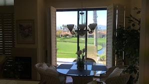 Ein Essbereich mit Korbstühlen und einem Tisch mit Glasplatte, einer Kristallleuchter und Blick auf einen Golfplatz durch offene Fenster.