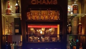 A bar area with a television screen displaying a news program, shelves of liquor bottles, and a refrigerator stocked with beverages. - The Chedy Home (Ariana)