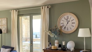 A room with a large wall clock, a wooden cabinet with various items, a blue sofa, and a view through a glass door to an outdoor area with tables. - les-pepites-de-Badou.fr present LA GRAVIERE -Classified 4 *- (Le Barroux)
