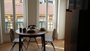 Ruokailutilassa on puinen pöytä ja tuolit, suuret ikkunat, joista on näkymä rakennuksiin, sekä jääkaappi.