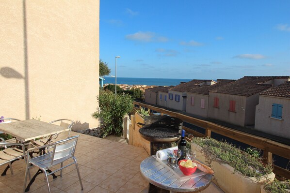 A patio with a table and chairs, a grill, and a view of the sea and buildings.