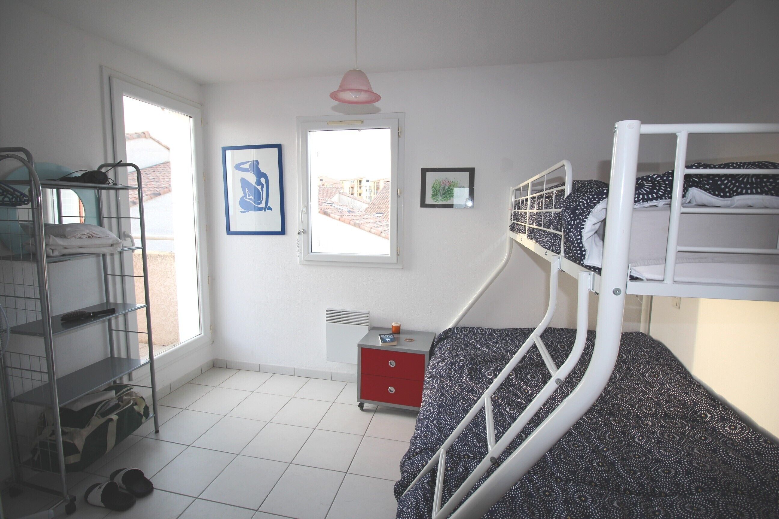 A dormitory room with bunk beds, a metal shelf unit, a small desk, and a window with a view of buildings.