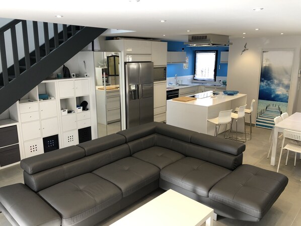 A modern kitchen with a central island, a dining area with white chairs, and a grey sectional sofa.