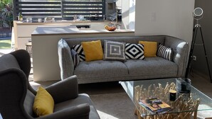 A modern living room with a grey sofa, yellow cushions, a glass coffee table, and a kitchen area with a large window.