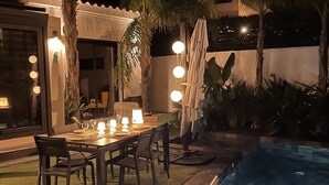 An outdoor dining area with a pool, surrounded by palm trees and a well-lit patio.
