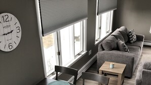 Ein modernes Wohnzimmer mit Essbereich, ausgestattet mit einem Holztisch mit blauen Untersetzern, einem grauen Sofa und einer Wanduhr.