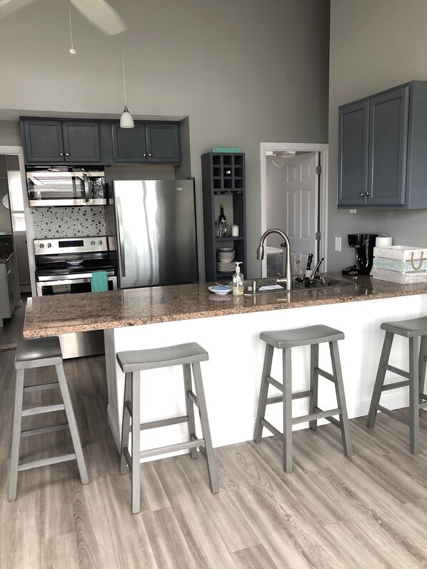 A modern kitchen with a granite countertop, stainless steel appliances, and a built-in oven. There are three bar stools and a small island with a sink.