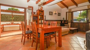 Un vaste espace de vie comprenant une table à manger en bois, des chaises, un canapé et une bibliothèque. Une fenêtre offre une vue sur la verdure environnante.