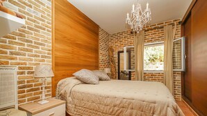 Une chambre avec un mur en briques, un panneau en bois, un lit recouvert d’une couette beige, une table de chevet avec une lampe, une commode et une fenêtre avec des rideaux.