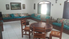 Una sala de estar con un sofá modular, una mesa redonda de madera y sillas. Hay cuadros enmarcados en la pared y una ventana con vistas.