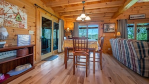 A wooden cabin interior with a dining table, chairs, a sofa, and a painting on the wall.