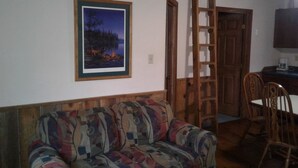 Ein gemütlicher Wohnbereich mit einem gemusterten Sofa, einem gerahmten Landschaftsbild, einer Holzleiter und einem Esstisch mit Stühlen.