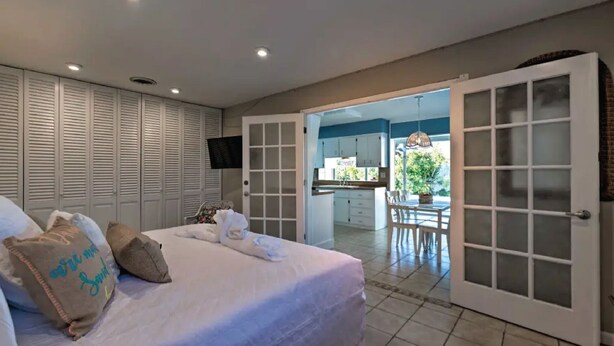 A bedroom with a bed, pillows, a TV, and a view of a kitchen and patio through a glass door. - Heated pool, large home across from the beach. Lots of amenities included. (Ormond Beach)