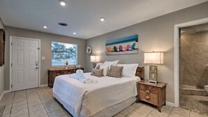A bedroom with a large bed, two bedside tables, a window with blinds, a painting above the bed, and a door. - Heated pool, large home across from the beach. Lots of amenities included. (Ormond Beach)