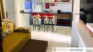 A compact kitchen with a microwave, stove, and refrigerator. There is a dining area with a small table and four red chairs. A sofa is placed in the living area. - Lacqua di Roma Hotel with Water Park and Flats complete with Kitchen (Caldas Novas)