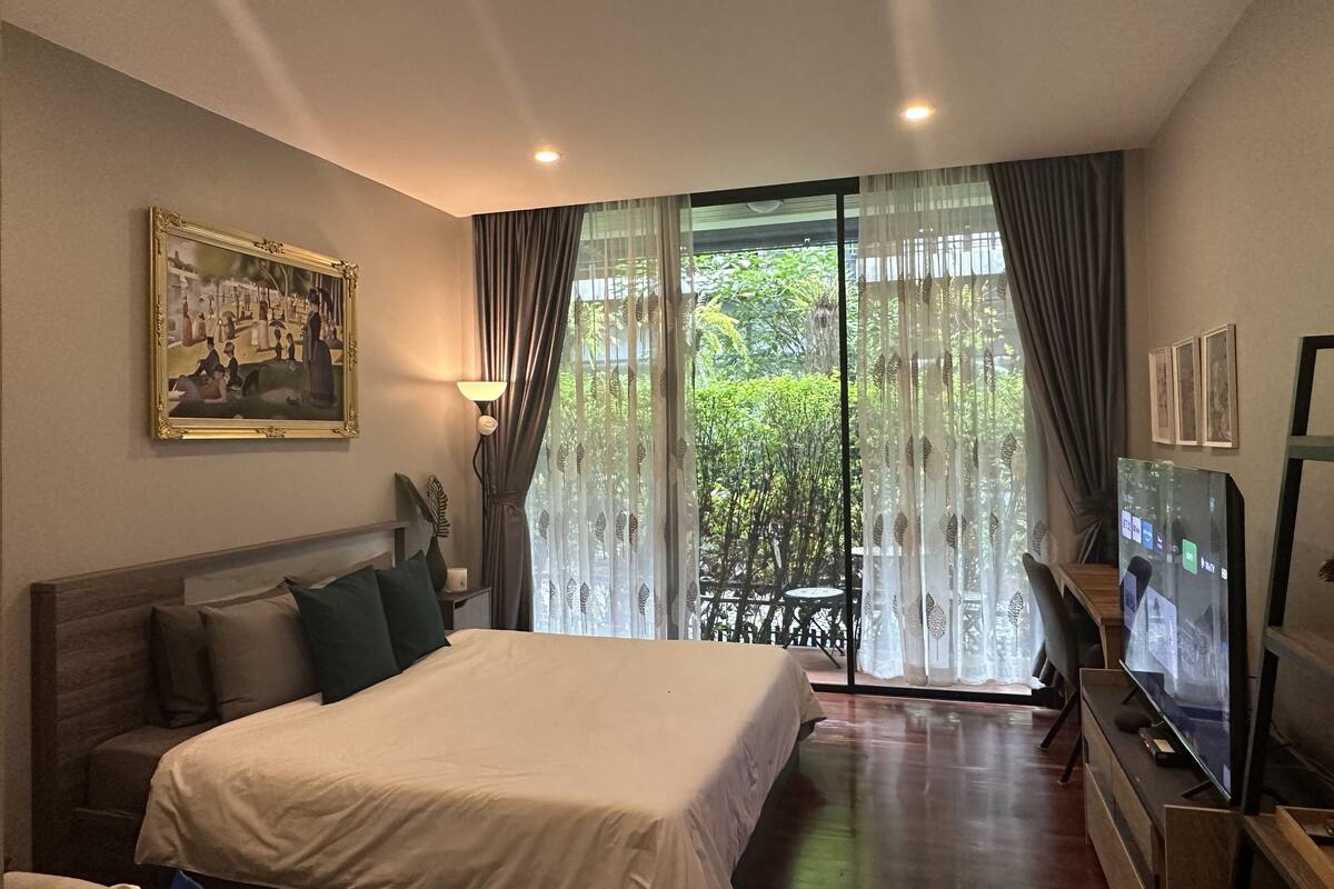A hotel room with a bed, a television, a painting, and a view of greenery through the window.
