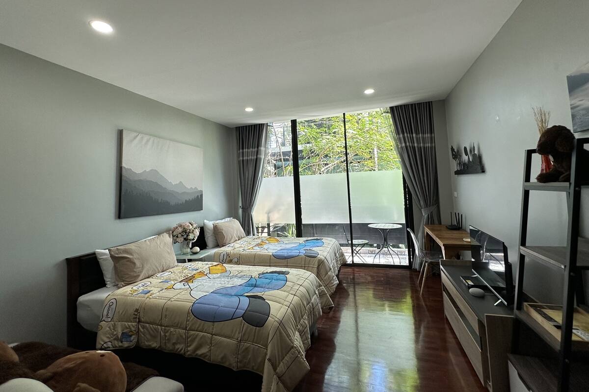 A modern bedroom with two beds, a large window, and a view of greenery outside.