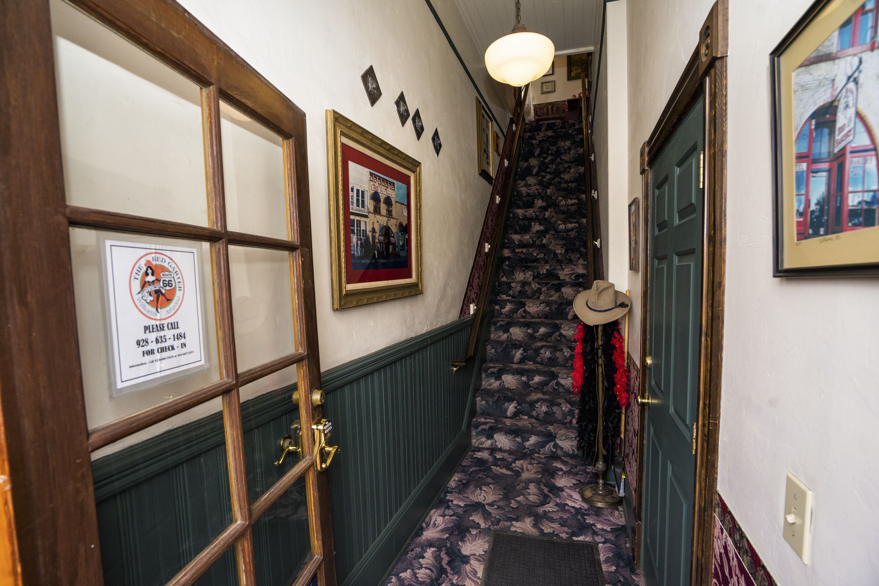 A hallway with a wooden door, a framed sign, a staircase with patterned carpet, and a door with a hat hanging on it.