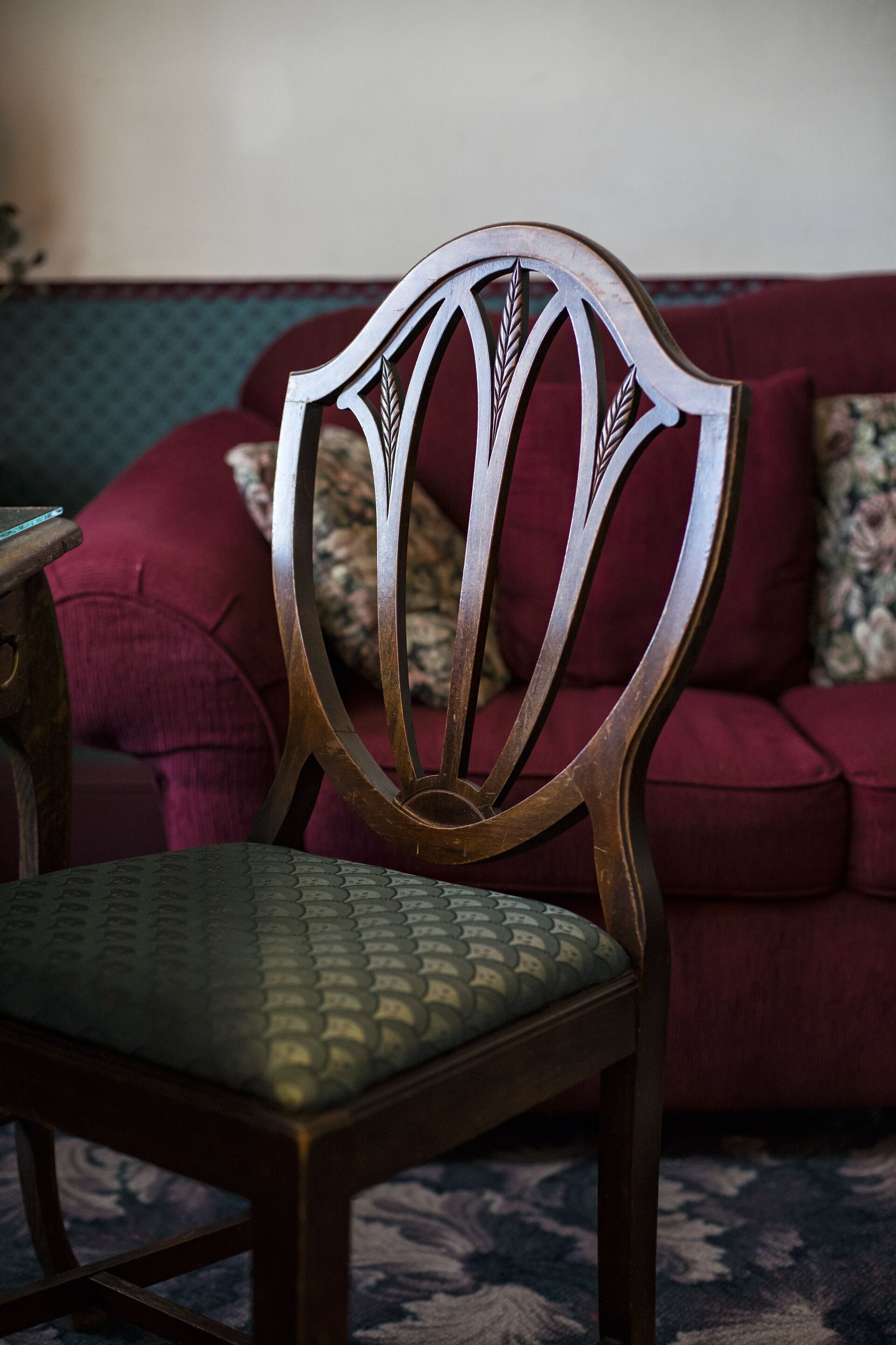 A wooden chair with a cushioned seat and backrest, positioned in front of a red sofa with patterned cushions.