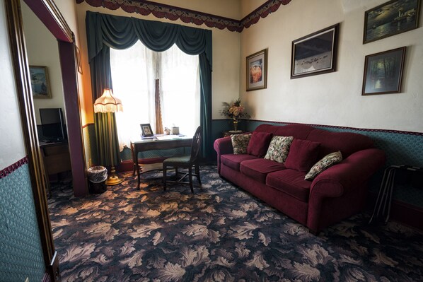 A room with a patterned carpet, a red sofa, a desk with a lamp, and framed pictures on the wall.