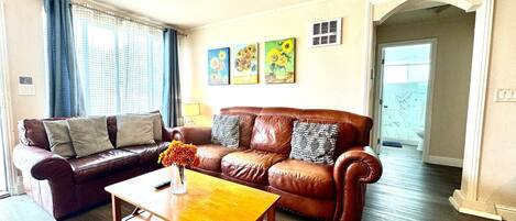 A living room with a brown leather sofa, a wooden coffee table, and a vase with flowers. There are three paintings on the wall and a window with curtains.