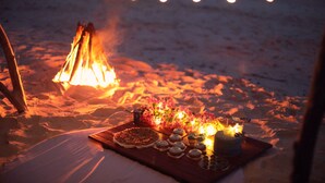 Een strandscène met een kampvuur, lichtjes en een picknickopstelling met eten en drinken.