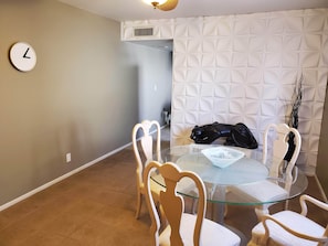 A dining area with a glass table and wooden chairs, a clock on the wall, and a textured white wall in the background.