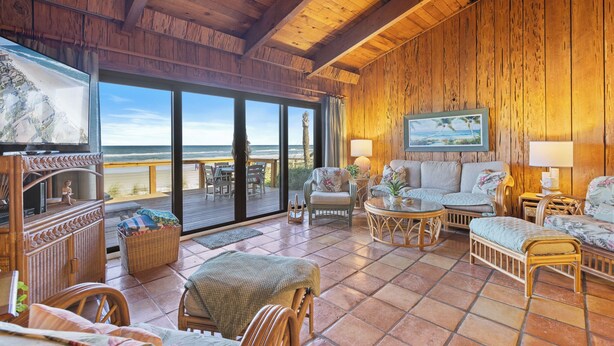 A spacious room with wooden walls and ceiling, large windows, a sofa, chairs, a table, and a television.