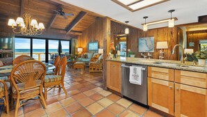 A spacious kitchen with wooden cabinets, a dishwasher, and a large island with a sink. There is a dining area with wicker chairs and a view of the ocean.