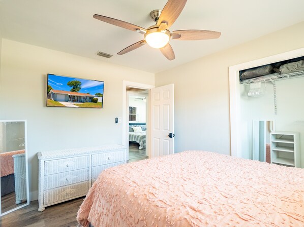 A bedroom with a ceiling fan, a bed with a pink blanket, a dresser, a mirror, and a door leading to another room. - Remodeled Home with a Pool, Covered Lanai, King Bed, 4 Bedrooms, and BBQ Grill! (Clearwater)