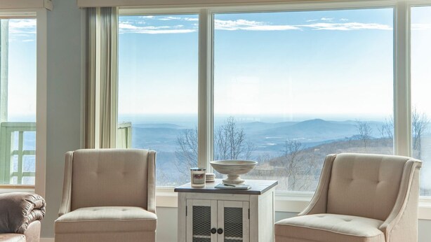 A room with two beige armchairs, a small table with a bowl and a container, and a large window offering a view of distant mountains. - WINTERGREEN RESORT HOME WITH BLUE RIDGE VIEWS (Wintergreen Resort)