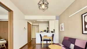 A modern living room with a wooden floor, a sofa, two small tables, a dining area with a table and chairs, and a kitchen in the background. - Luana Lea Karate Kaikan Mae (Tomigusuku)