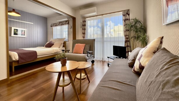 A modern, well-lit room with a sofa, a bed, a TV, and a small table. - Luana Lea Karate Kaikan Mae (Tomigusuku)
