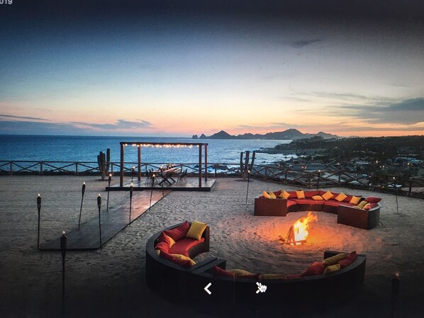 A beachside seating area with a fire pit, a covered dining area, and a view of the ocean and distant mountains.