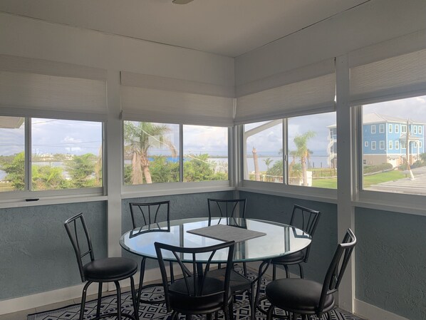A room with a glass table and chairs, large windows with a view of the sea and buildings, and a patterned rug on the floor. - Dolphins, Sunsets & Waterfront Living – Heated Pool + Dock & Kayaks Near Beaches (Englewood)