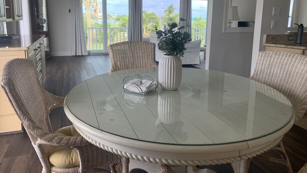 A dining area with a round glass table, wicker chairs, a vase with greenery, and a view of the outdoors through large windows. - Dolphins, Sunsets & Waterfront Living – Heated Pool + Dock & Kayaks Near Beaches (Englewood)